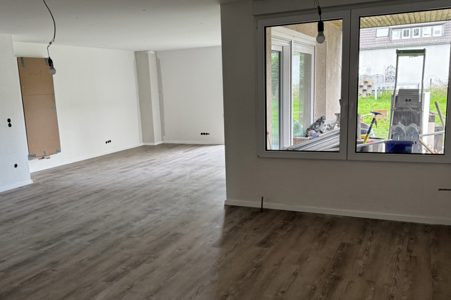 Leeres Wohnzimmer mit Blick durch Fenster auf überdachte Terrasse, Boden in Holzoptik, HolzLand Beese Unna